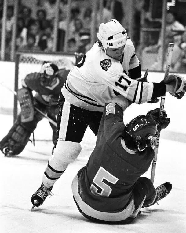 A hockey player in a white uniform and helmet checks an opposing player in a dark uniform who is falling to the ice. A goalie stands ready in the background near the goal.