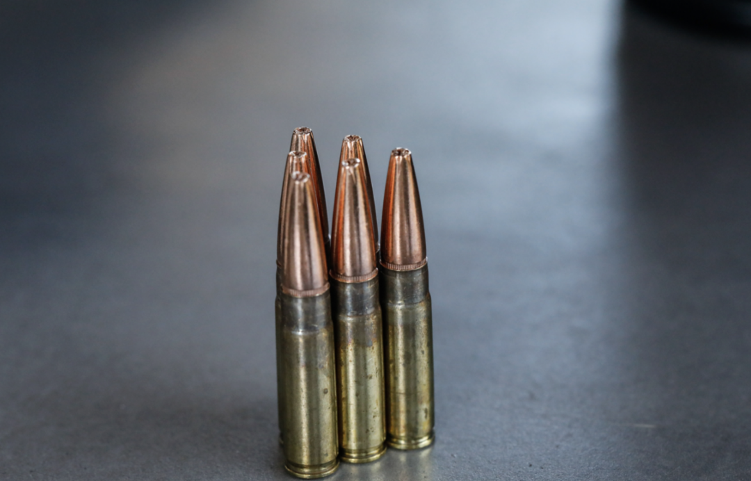 Three rifle bullets with brass casings and pointed copper tips are standing upright on a gray surface, closely aligned side by side.