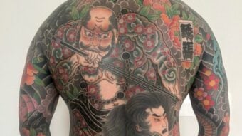 A person&rsquo;s back covered with a colorful Japanese-style tattoo, featuring two fierce warrior figures surrounded by intricate floral patterns and traditional motifs.