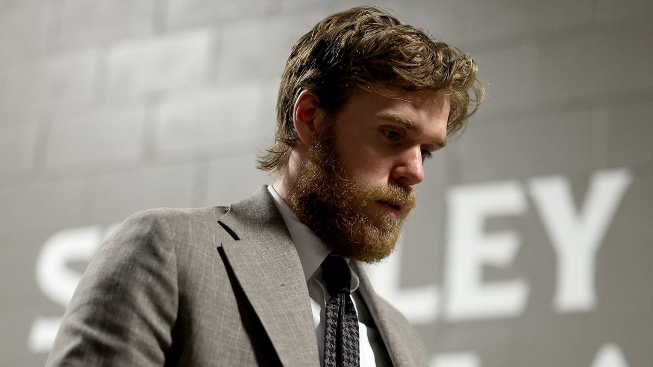 A man with a brown beard and mustache, wearing a gray suit and tie, looks downward with a serious expression. The background is a gray wall with partially visible white text.