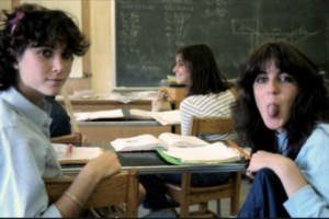 Two teenage girls sit at desks in a classroom; one looks at the camera while the other playfully sticks out her tongue. A chalkboard and two more students are visible in the background.