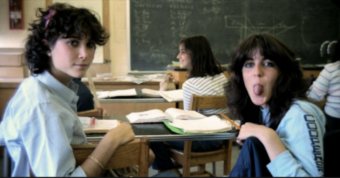 Two teenage girls sit at desks in a classroom; one looks at the camera while the other playfully sticks out her tongue. A chalkboard and two more students are visible in the background.