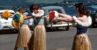 Three women in grass skirts and flower leis perform a hula dance with colorful pom-poms in a parking lot, with classic cars and a scenic landscape in the background.