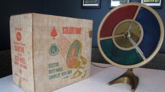 A vintage electric Colortone Roto-Wheel with red, green, yellow, and blue panels sits beside its original box on a table. Framed posters are visible on the wall in the background.
