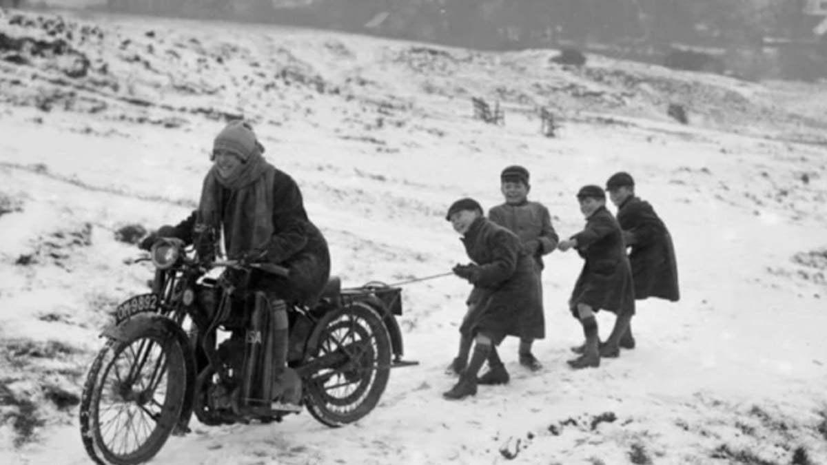 A person rides a motorcycle through a snowy field while four children, bundled in coats, hold a rope tied to the back and are pulled along behind. It appears to be a winter scene from the past.