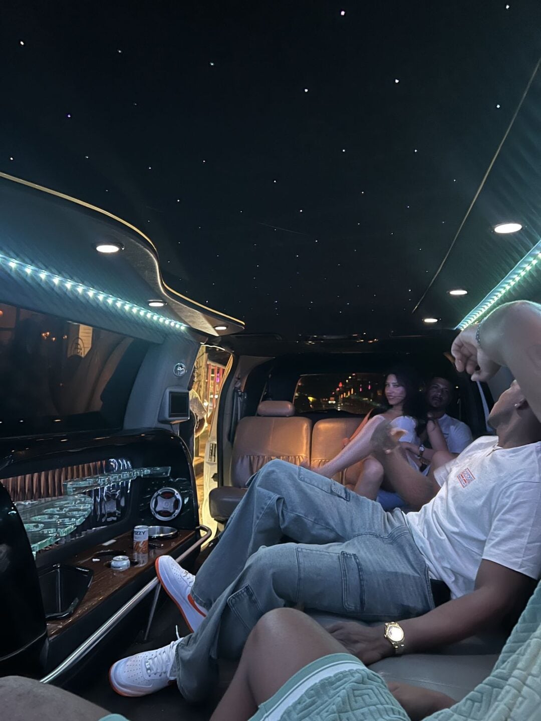 Several people relax inside a luxurious limousine with leather seats, blue LED lighting, and a starry ceiling effect. Some chat while one person stretches out, showing white sneakers and a gold watch.