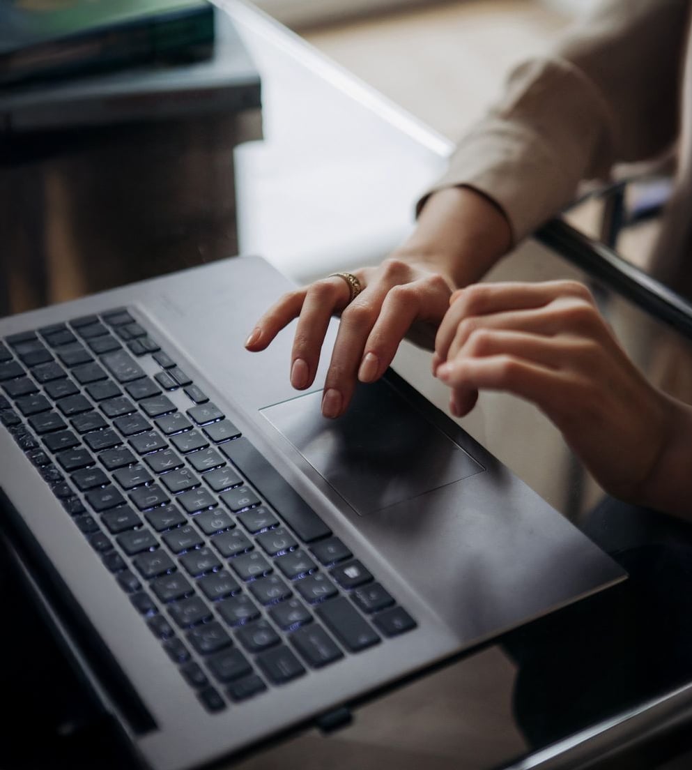 A person using a laptop, with one hand on the keyboard and the other on the touchpad, sitting at a glass desk. The person is wearing a beige long-sleeve shirt and a ring on one finger.