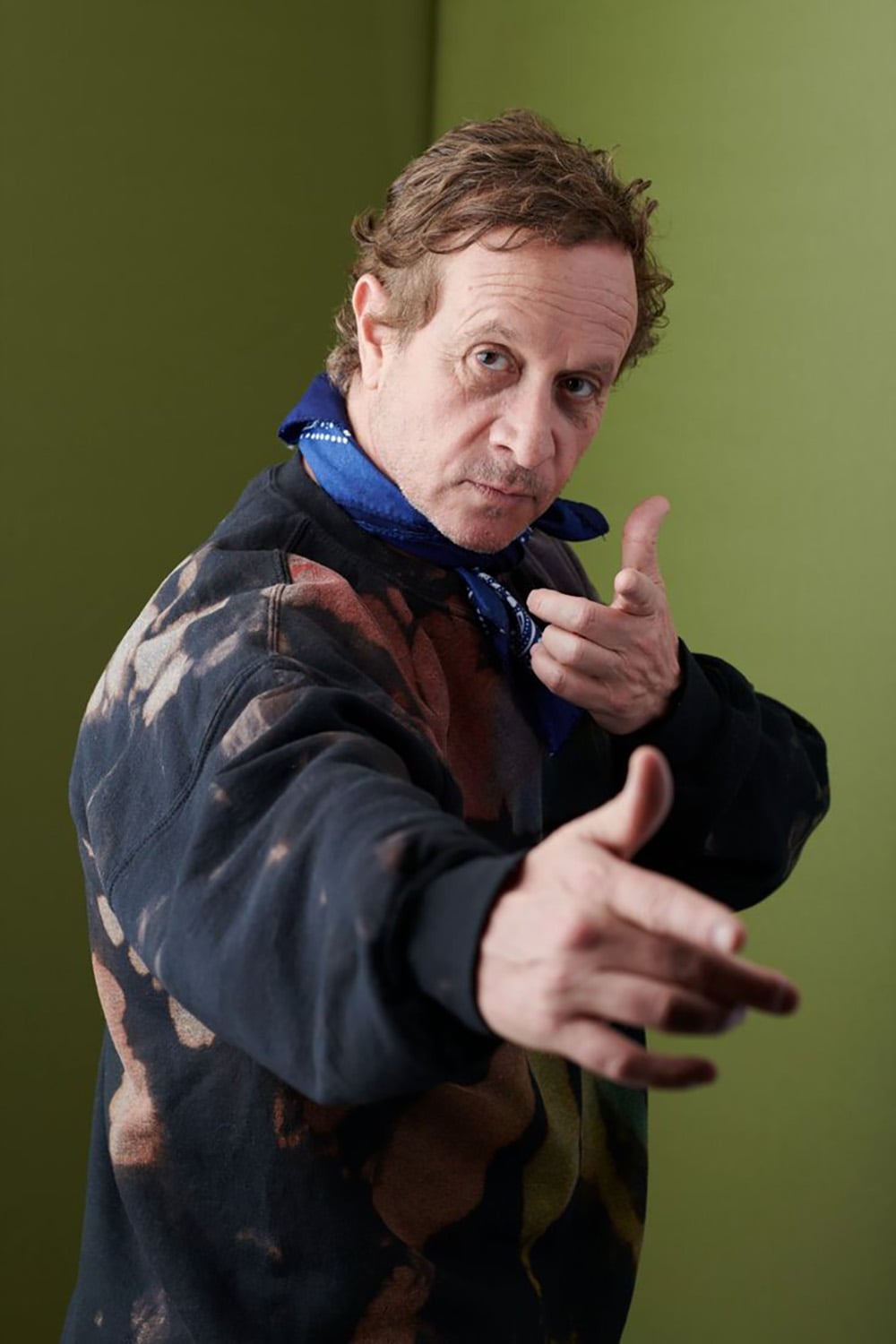 A man with short brown hair poses against a green background, pointing his fingers forward as if mimicking a finger gun. He wears a dark tie-dye sweatshirt and a blue bandana around his neck.