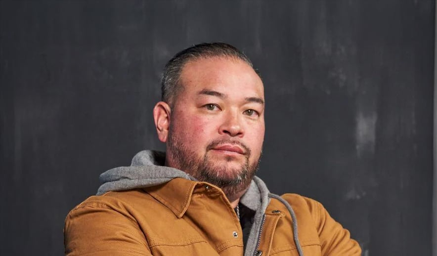 A man with short hair and a beard, wearing a brown jacket over a gray hoodie, stands in front of a dark, textured background with a neutral expression.
