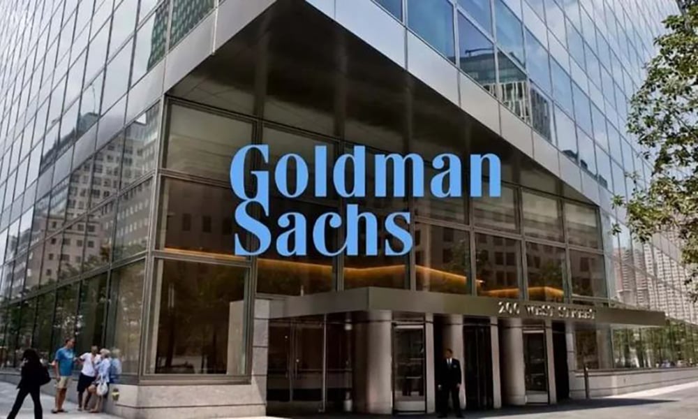 The glass entrance of a modern office building displays a large "Goldman Sachs" sign. Several people are walking outside, and the building reflects nearby city structures.
