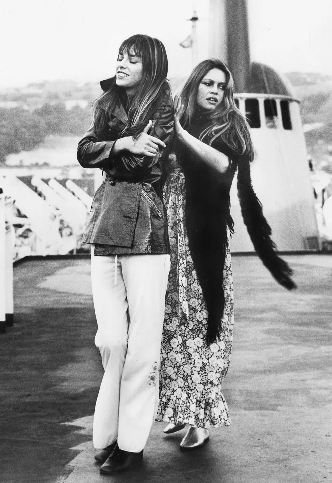 Two women in 1970s fashion stand on the deck of a ship; one wears a jacket and wide-legged pants, the other a long floral dress and feather boa. They smile and interact playfully with each other.