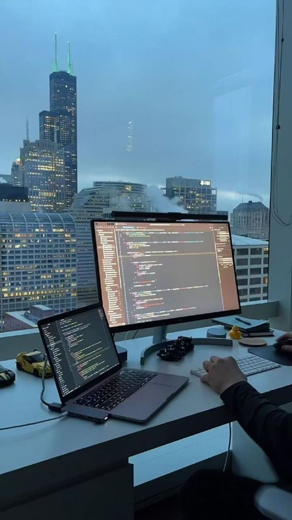 A person works at a desk with a laptop and large monitor displaying code. The workspace is next to a window overlooking a city skyline with tall buildings at dusk or dawn. Small desk items are also visible.