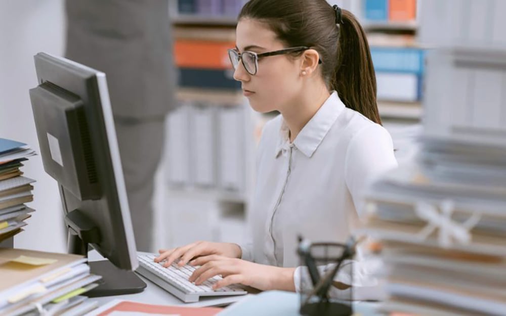 A woman wearing glasses and a white blouse is working at a computer in an office, surrounded by stacks of documents and folders. She appears focused on the screen.