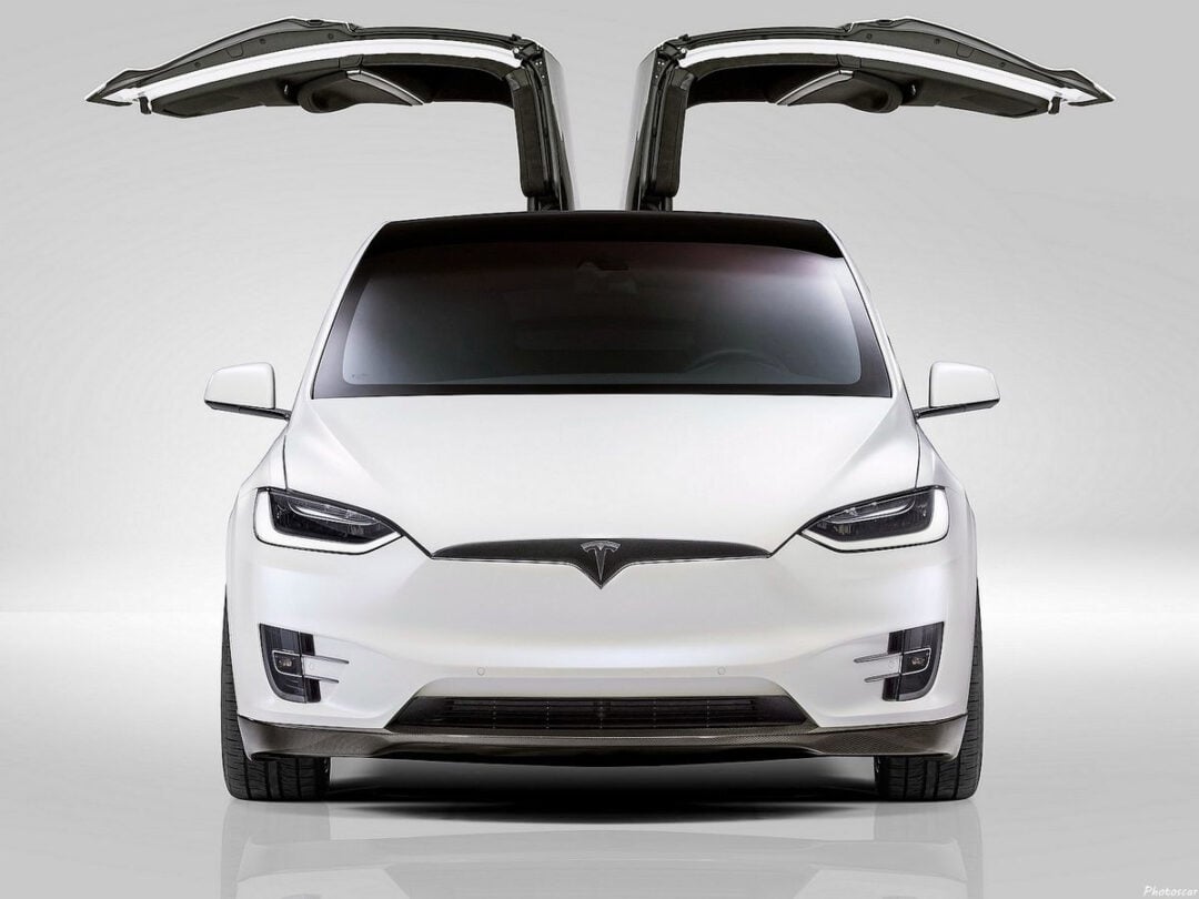 A white Tesla Model X electric SUV is shown from the front with its falcon wing doors open upwards, set against a plain white background.