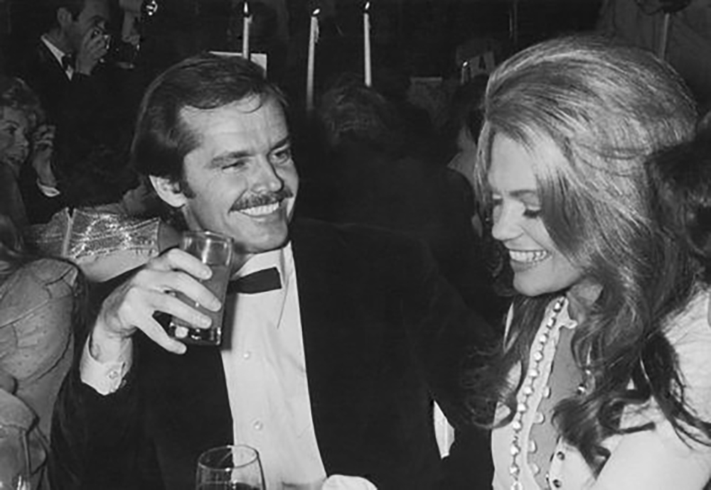 A man in a tuxedo smiles and raises a drink while sitting next to a woman with long hair and a pearl necklace at a lively party. Both appear happy and are engaged in conversation.