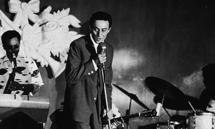 A man in a suit sings into a microphone on stage, accompanied by a saxophonist and a drummer, with a large decorative backdrop behind them. The image is in black and white.