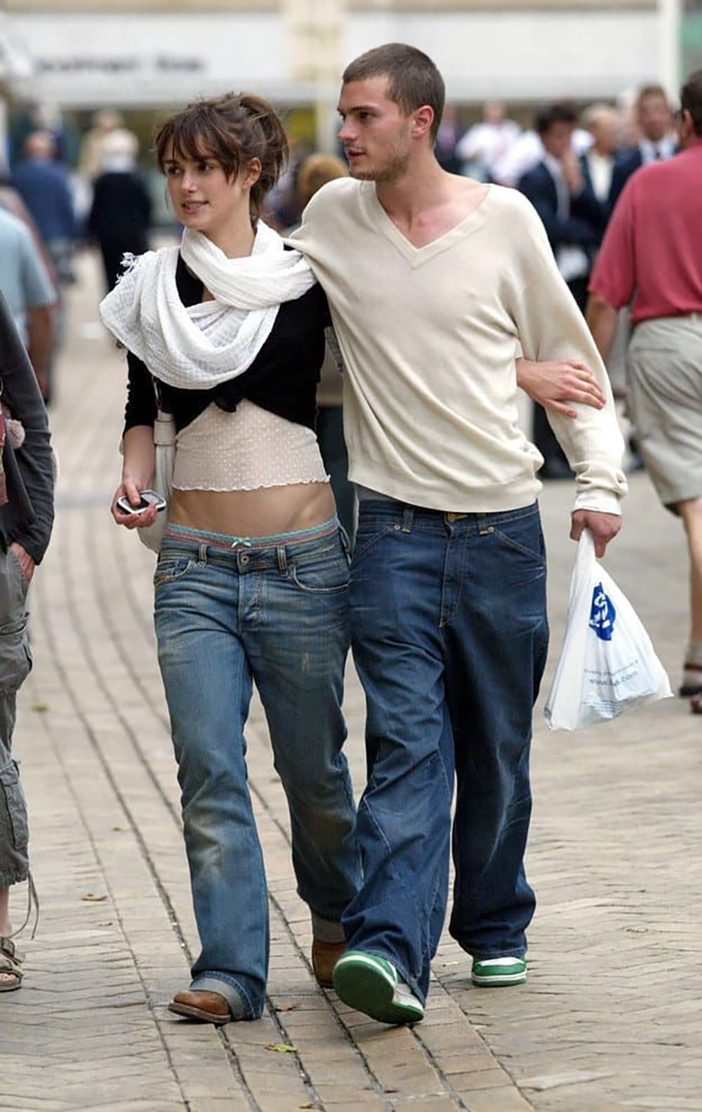 A young woman and man walk arm in arm down a street. The woman wears low-rise jeans, a crop top, and a white scarf, while the man wears loose jeans and a light sweater, holding a plastic bag. People walk in the background.