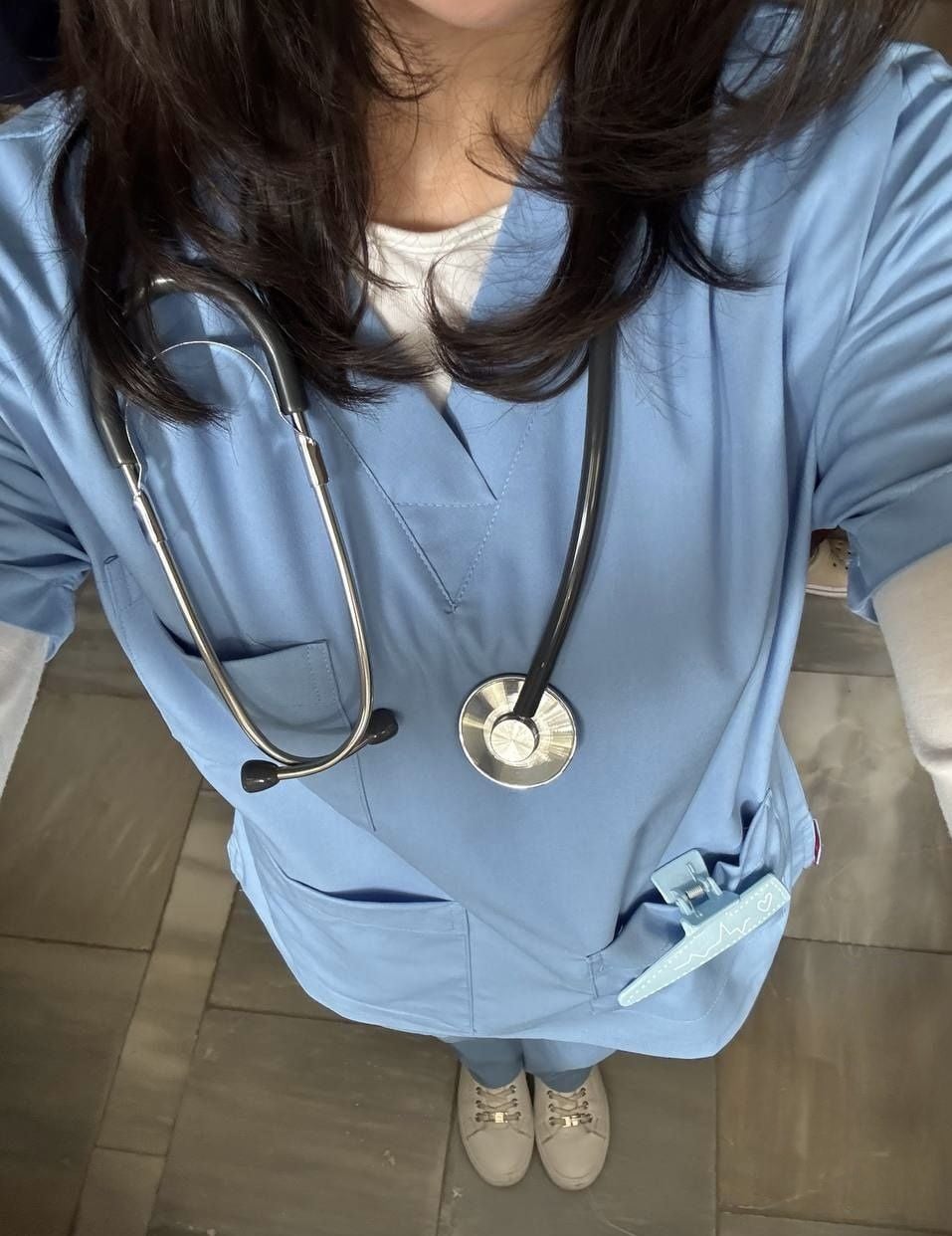 A person wearing blue medical scrubs with a stethoscope around their neck stands on a tiled floor. Only the lower half of their face is visible. A pen and ID badge are in the pocket.