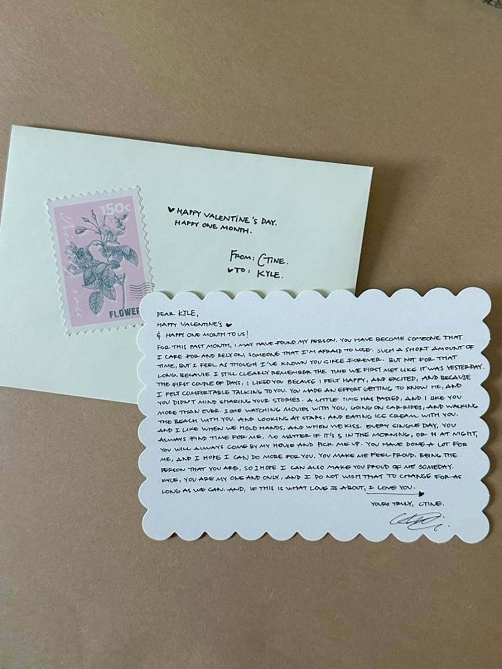 A handwritten Valentine's Day card with scalloped edges sits on a table next to an envelope with a pink flower stamp. The card contains a personal love note addressed to Kyle from Cathe.