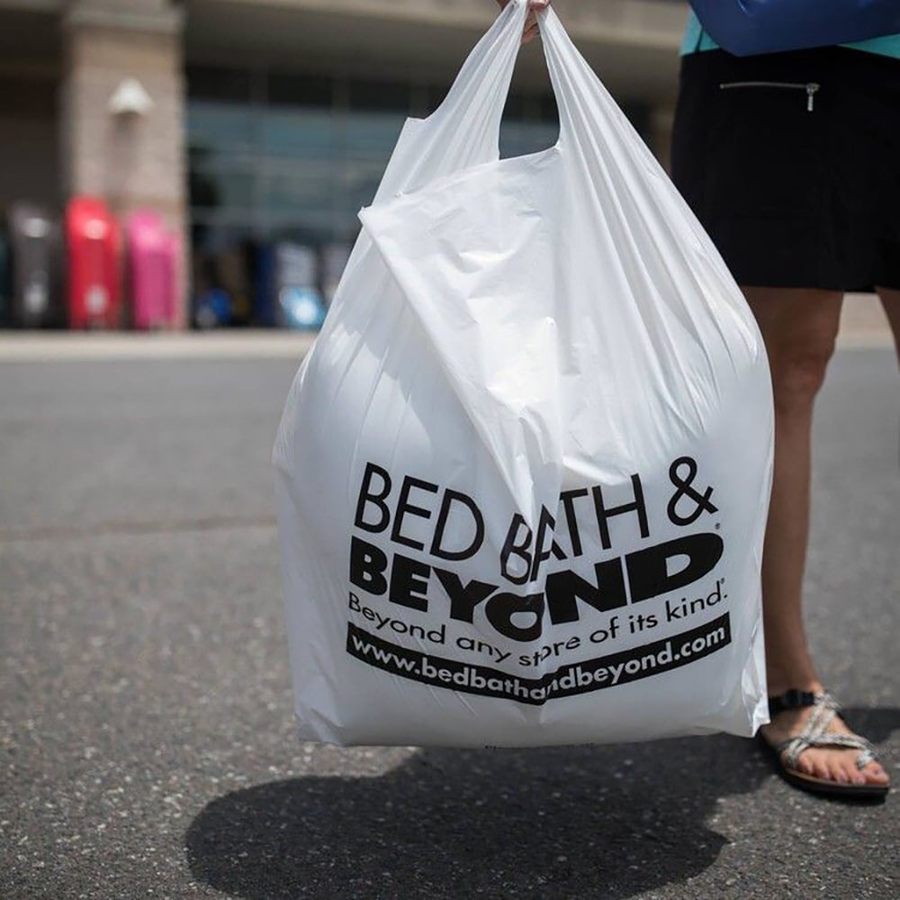 A person holds a large white Bed Bath & Beyond shopping bag outside, with the store’s logo and website clearly visible on the bag. The background shows pavement and blurred storefronts.
