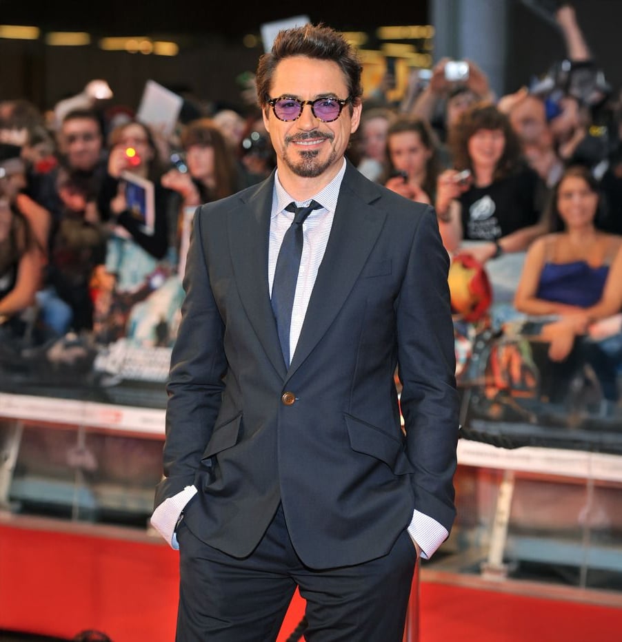 A man in a dark suit and sunglasses smiles with his hands in his pockets on a red carpet, with a cheering crowd and cameras in the background.