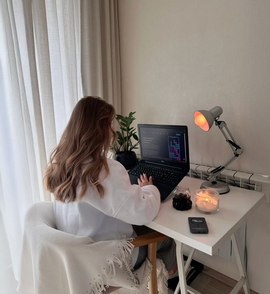 A person with long wavy hair sits at a white desk using a laptop. On the desk are a potted plant, a lit lamp, candles, a glass of a dark drink, and a smartphone. Light streams in through sheer curtains.