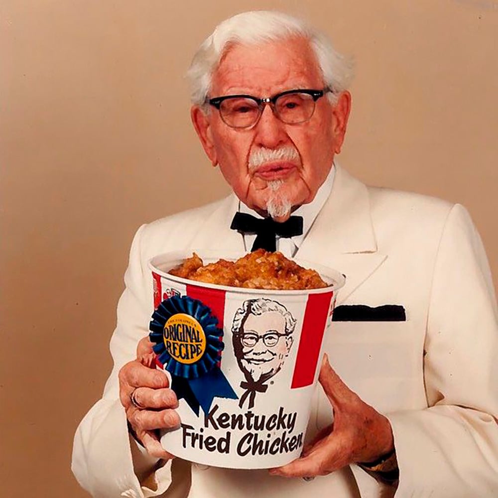 An elderly man with white hair, glasses, and a white suit holds a large bucket of Kentucky Fried Chicken labeled “Original Recipe,” with an image of his face on the bucket.