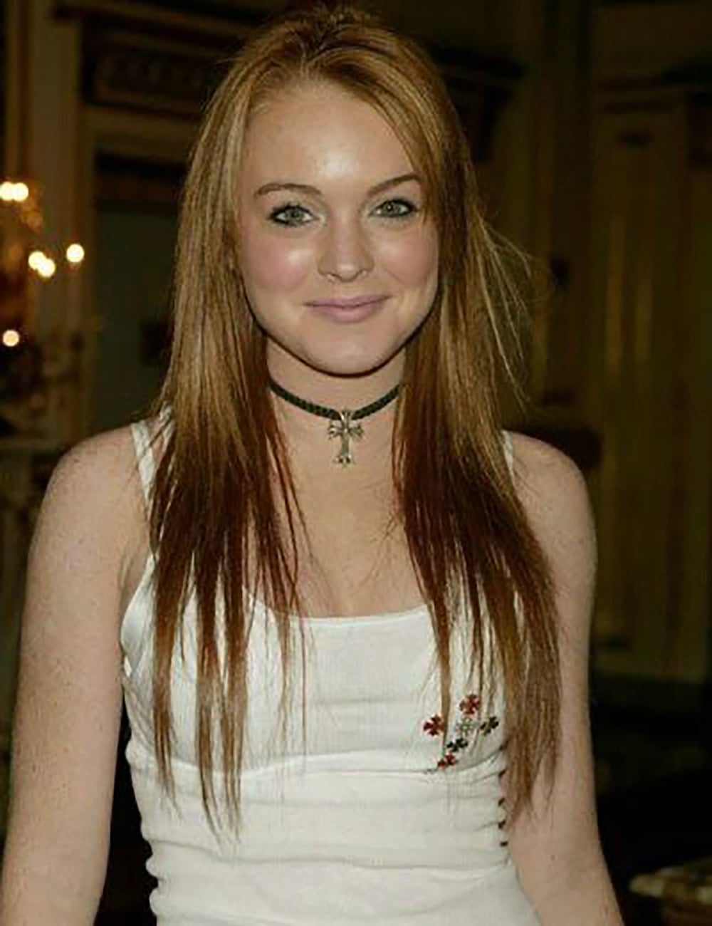 A young woman with long straight red hair and fair skin smiles at the camera. She wears a white tank top with small floral embroidery and a black choker necklace. The background is softly lit indoors.