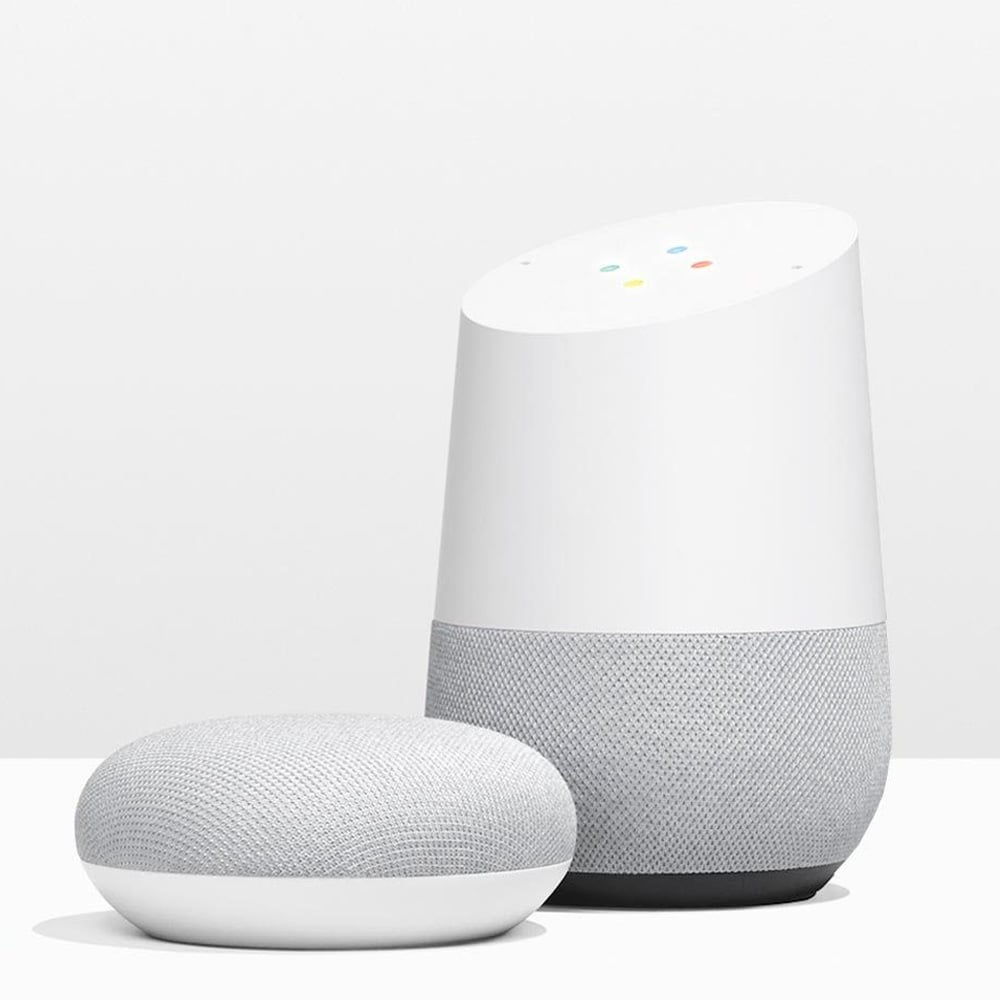 A white Google Home smart speaker stands next to a smaller, round Google Home Mini, both featuring a minimalist design with a gray fabric base and white top.