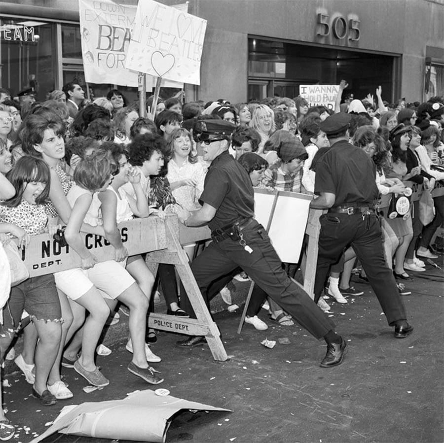 A black-and-white photo of excited young fans crowded behind a police barricade, holding Beatles signs, as two police officers try to hold them back outside a building with the number 505.