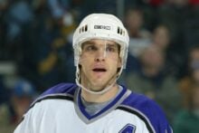 A male ice hockey player wearing a white helmet and a white jersey with purple and black accents, featuring an "A" patch on his chest, stands on the ice during a game. The background shows a blurred crowd.