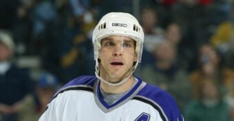 A male ice hockey player wearing a white helmet and a white jersey with purple and black accents, featuring an "A" patch on his chest, stands on the ice during a game. The background shows a blurred crowd.