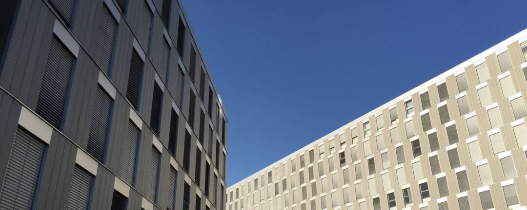 Two modern, multi-story buildings with grid-like window patterns rise on either side of the image, creating a narrow gap between them, with a clear blue sky visible above.