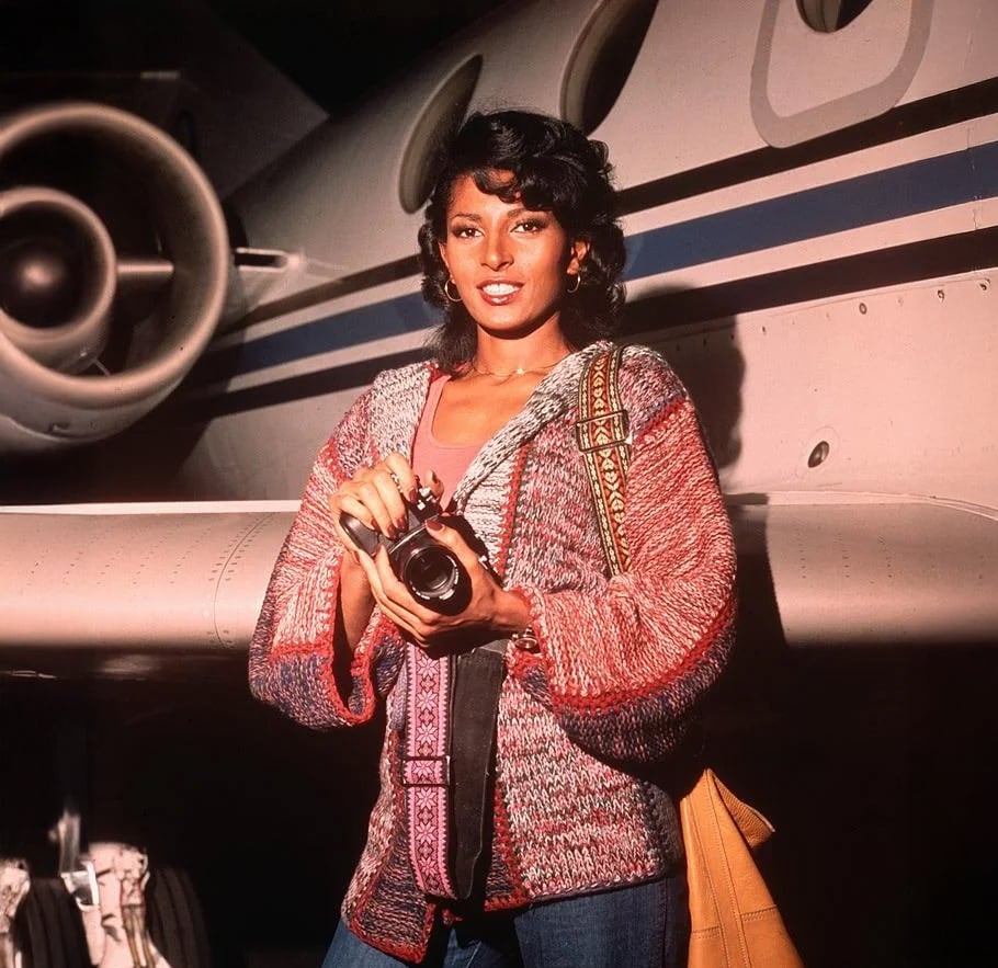 A woman is standing in front of a private jet, smiling and holding a camera. She wears a red and gray knit sweater and carries a tan shoulder bag, with the plane’s engine and fuselage visible behind her.