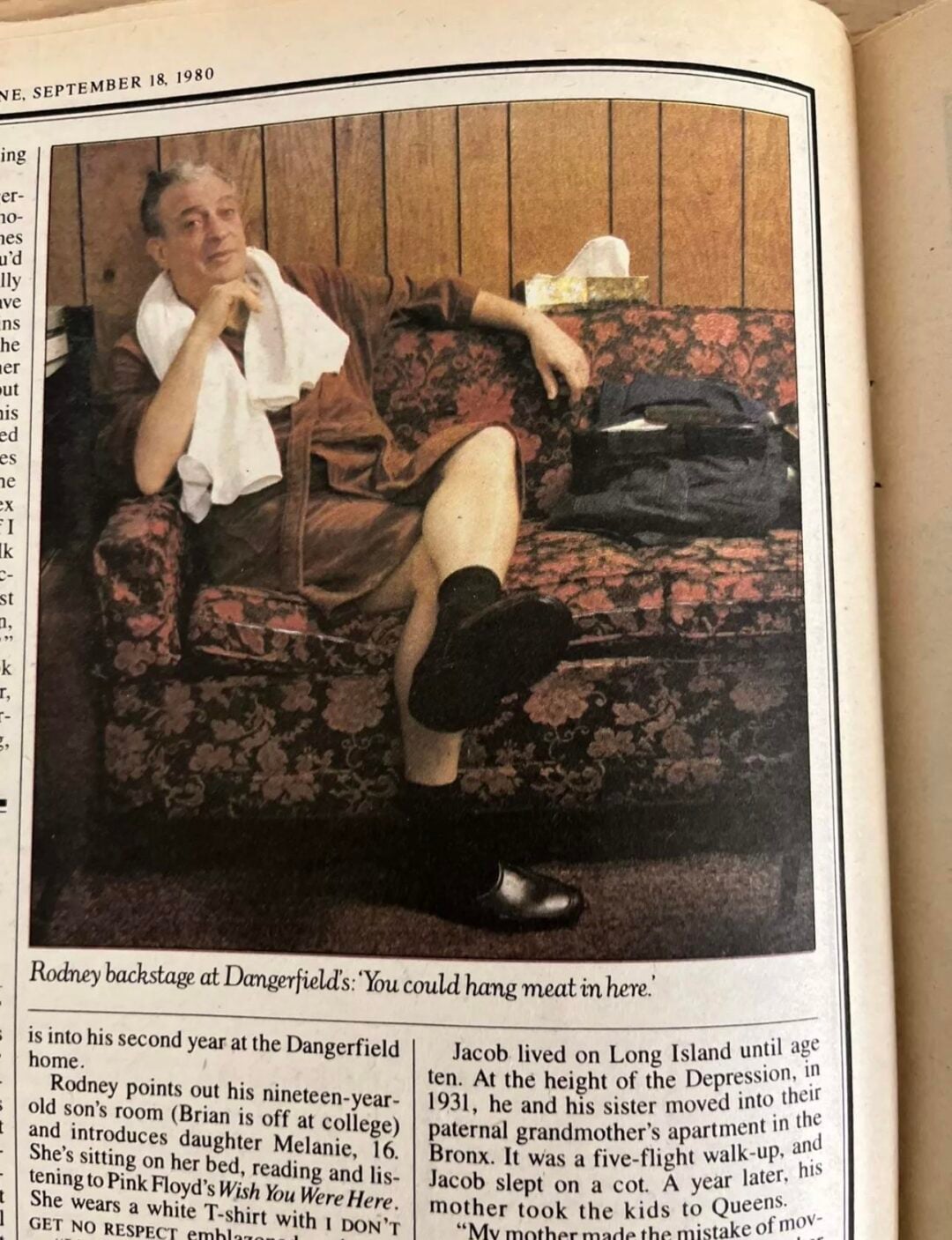 An older man sits on a floral couch with one leg crossed, wearing a white shirt, brown shorts, and black shoes. He has a towel draped over his shoulder and appears relaxed backstage, with a drink in hand.