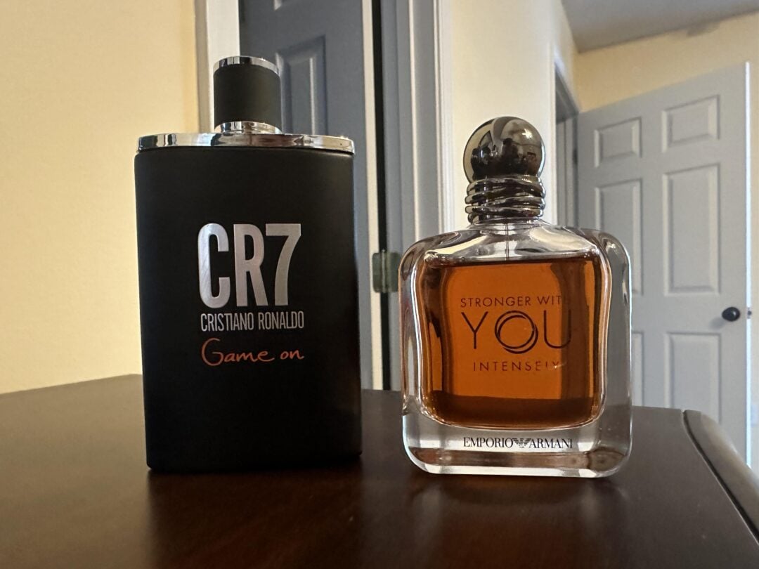 Two perfume bottles on a wooden surface: one black bottle labeled "CR7 Cristiano Ronaldo Game On," and one clear bottle labeled "Stronger With You Intensely Emporio Armani." Background shows open doors.