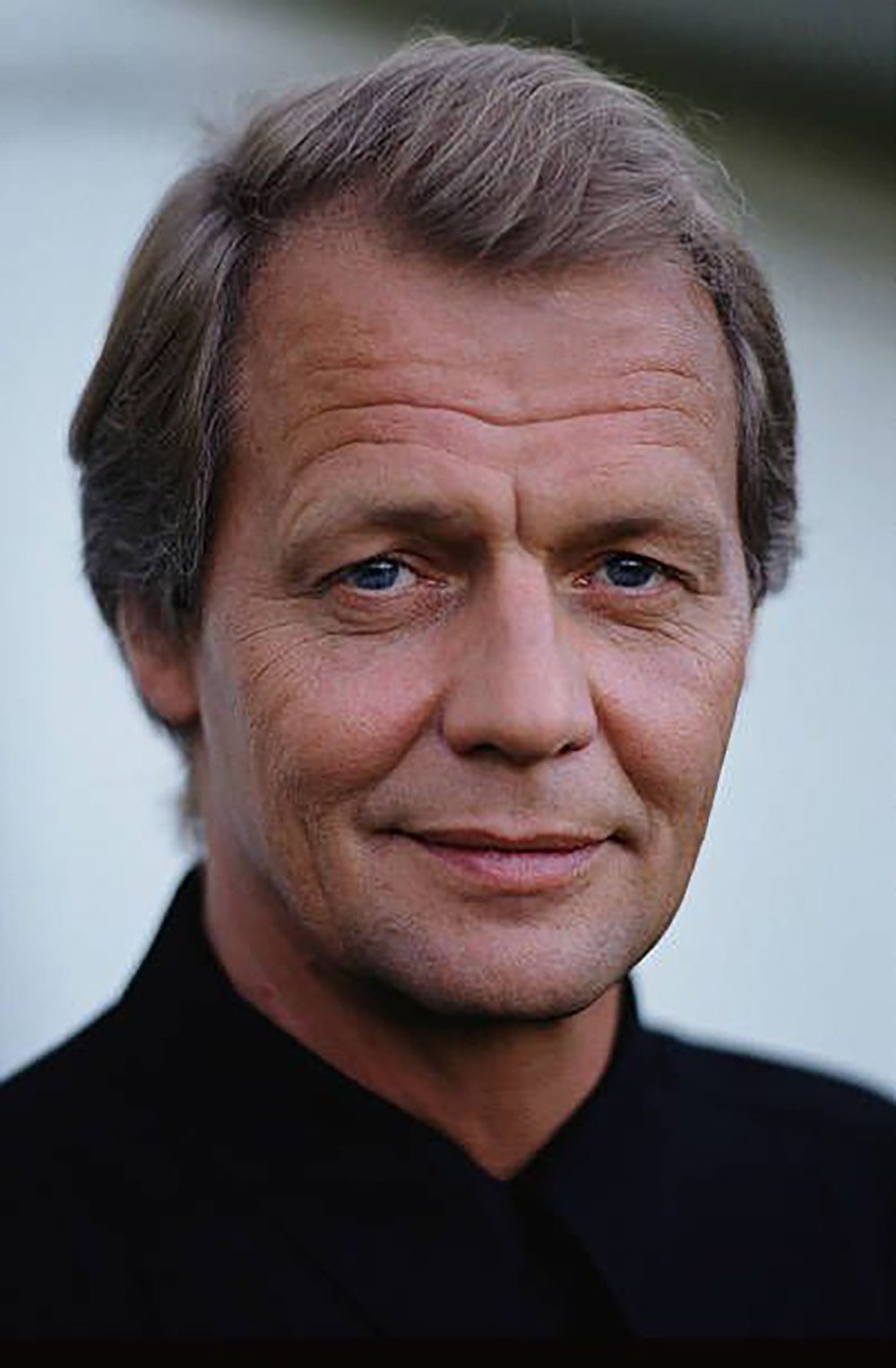 A middle-aged man with light brown hair and blue eyes, wearing a black shirt, looks directly at the camera with a neutral expression. The background is softly blurred.