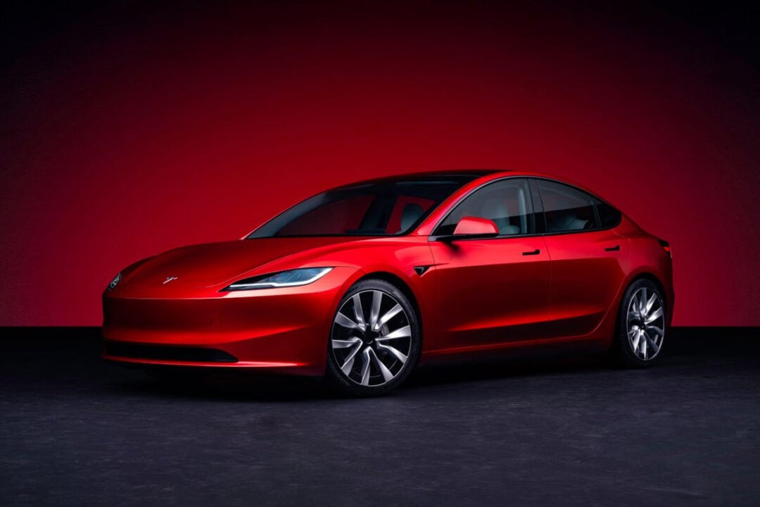 A sleek red Tesla sedan is parked in front of a gradient red background, showcased at an angle to highlight its modern design and aerodynamic lines.
