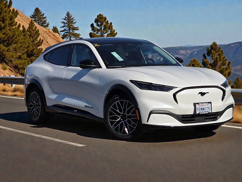 A white Ford Mustang Mach-E electric SUV drives on a mountain road lined with trees, under a clear blue sky.
