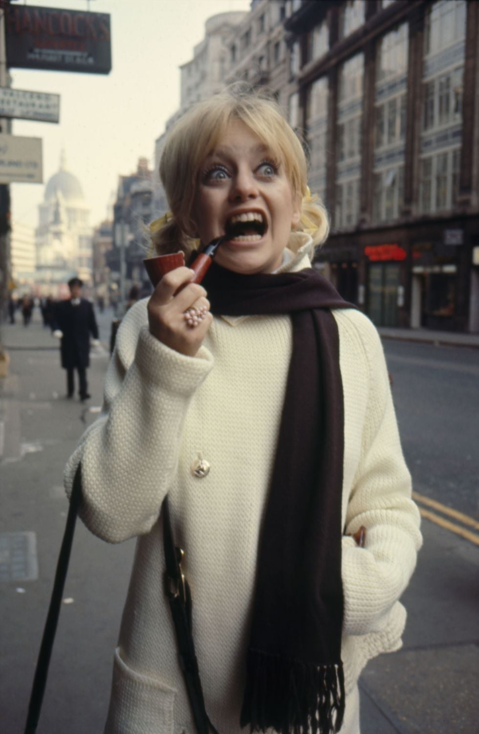 A woman with blonde hair in pigtails, wearing a white coat and dark scarf, stands on a city street with buildings behind her, holding a pipe and making a surprised or playful expression.