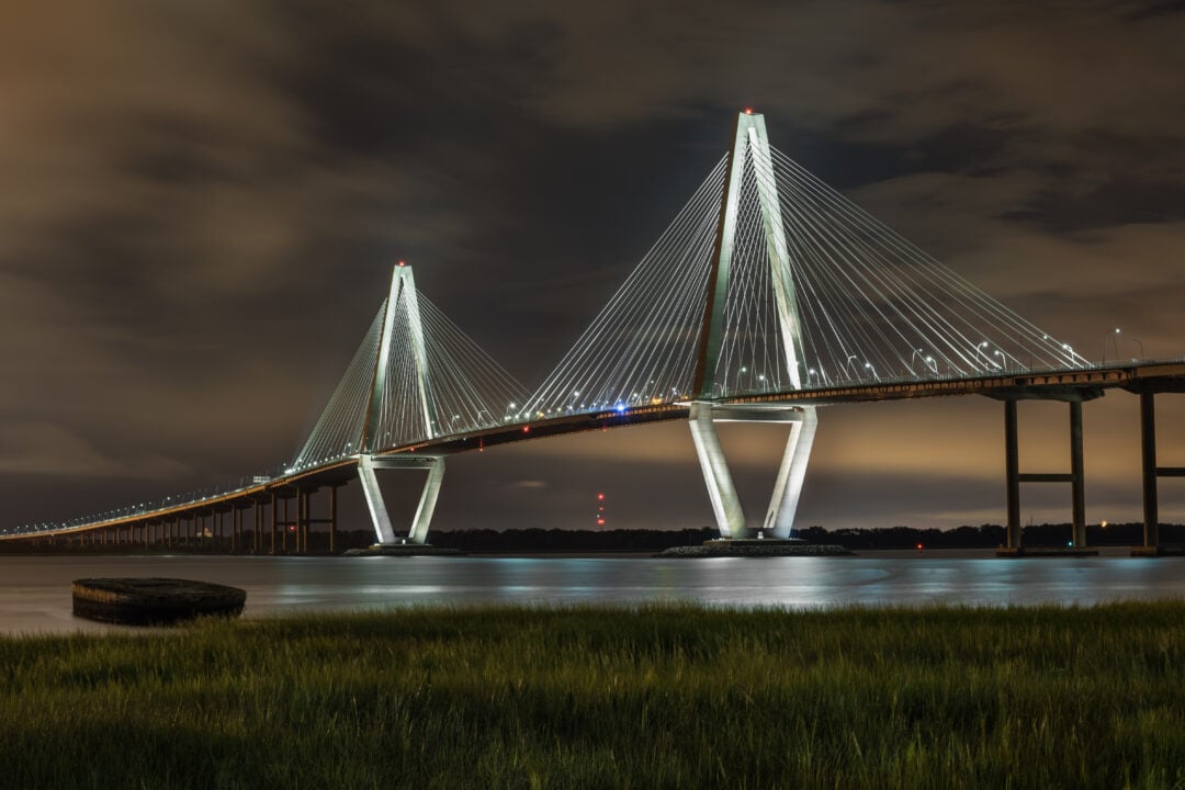 A modern, illuminated cable-stayed bridge spans across a calm river at night, with reflections on the water and grassy marsh in the foreground under a cloudy sky.