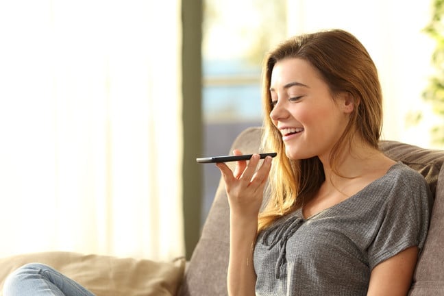 A young woman sits on a couch, smiling as she holds a smartphone in front of her mouth, speaking into it. Bright, natural light fills the room, creating a relaxed, comfortable atmosphere.