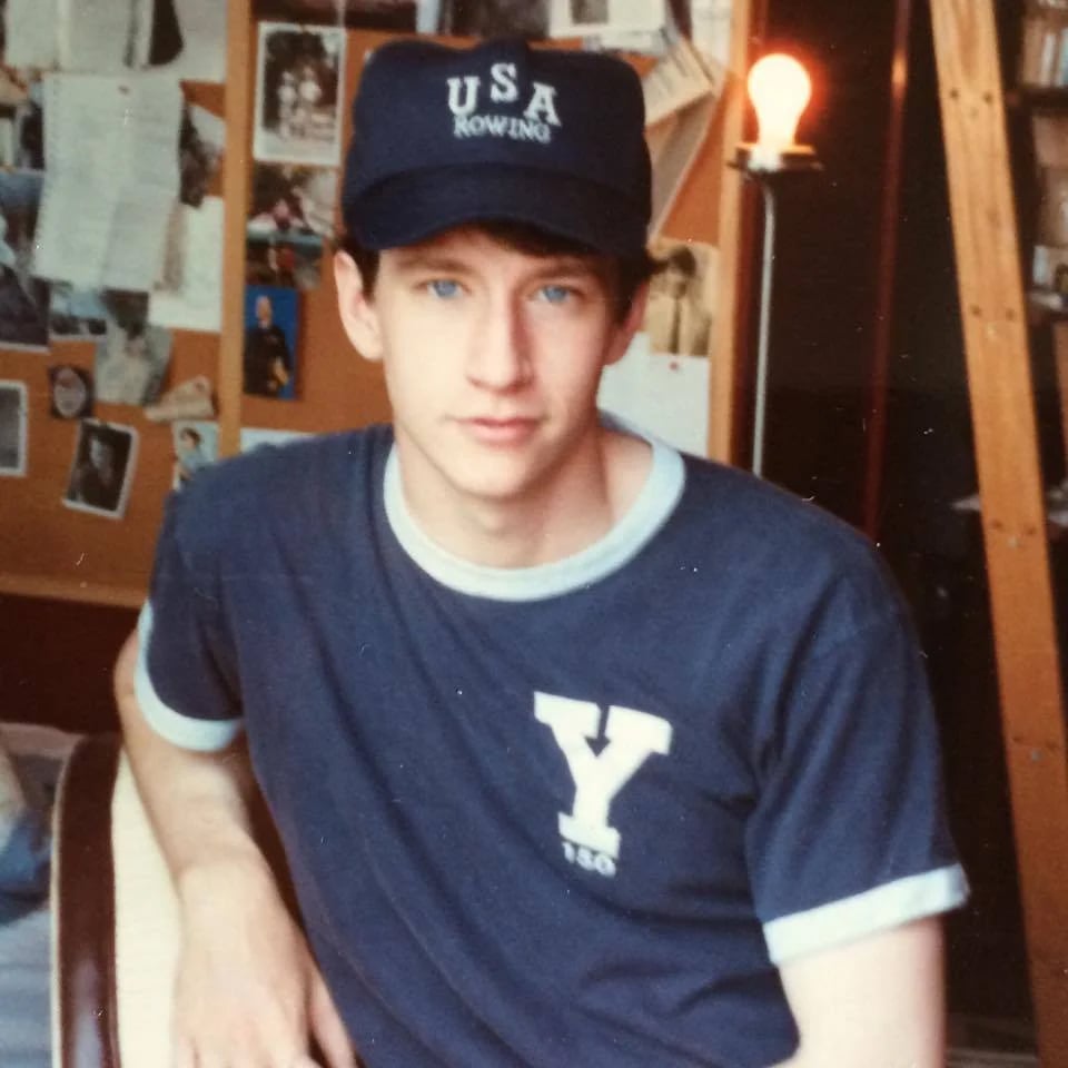 A young man with blue eyes wears a navy blue "USA Rowing" cap and a Yale t-shirt, sitting indoors. Behind him is a bulletin board with photos and papers, and a lit lamp on a shelf.