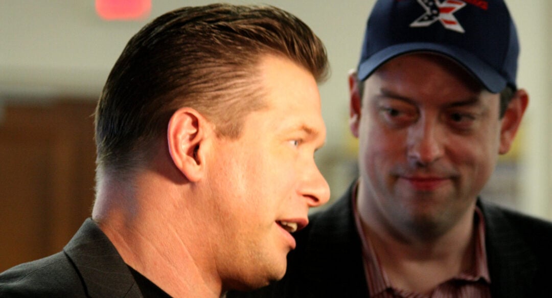 Two men indoors, one in the foreground speaking, wearing a black jacket and black shirt, and the other in the background wearing a striped shirt, dark jacket, and a blue baseball cap, looking at the first man.