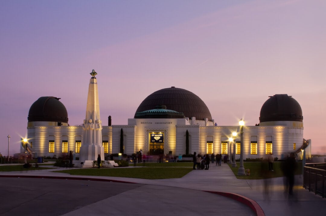 The Griffith Observatory is illuminated at dusk with a purple sky. The domed building features glowing windows and an art deco façade, with the Astronomers Monument standing prominently in front. Several people are visible outside.