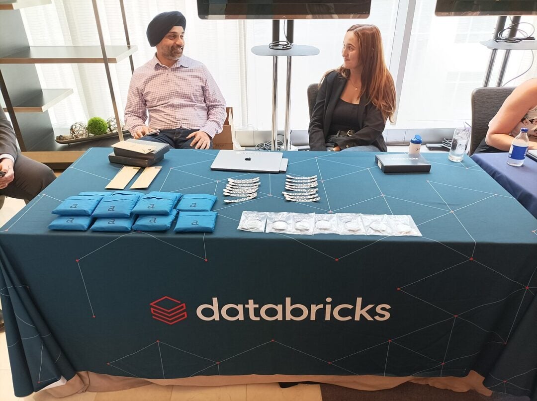 Two people sit behind a table covered with a blue Databricks tablecloth, displaying branded items, water bottles, and tech accessories at what appears to be a conference or expo.