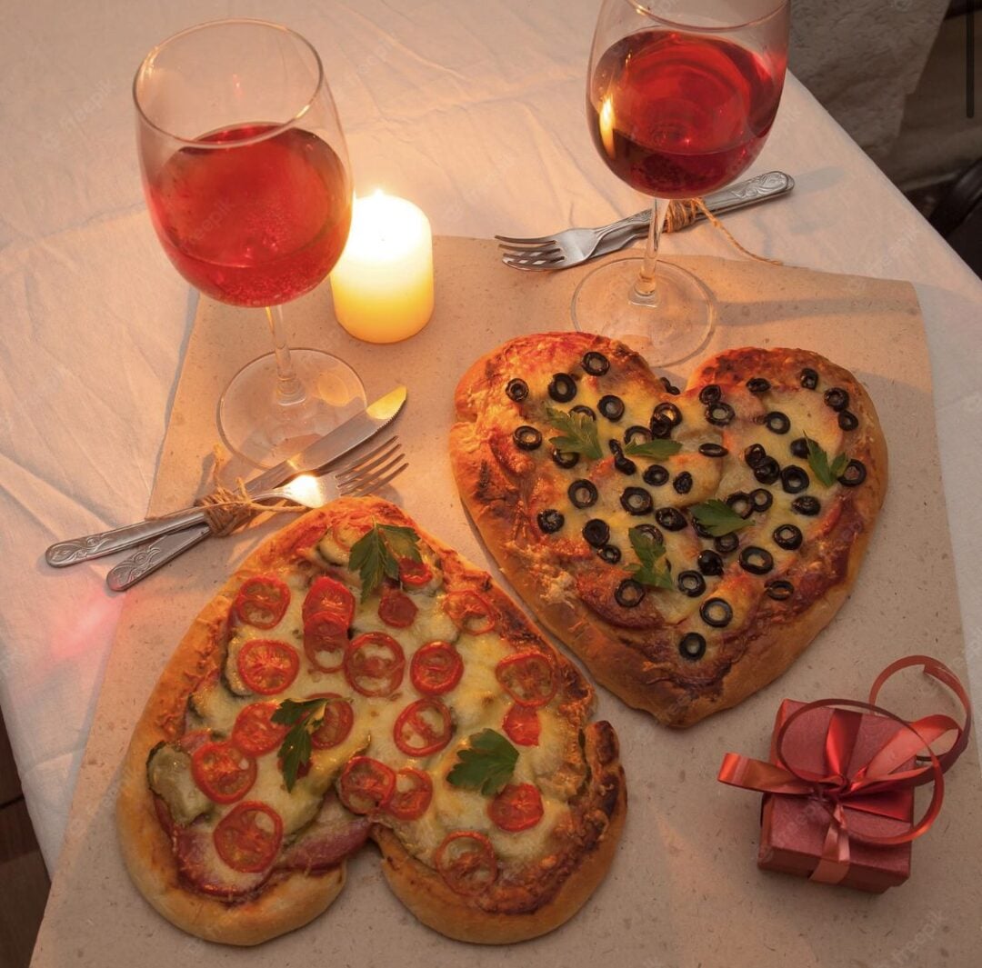 Two heart-shaped pizzas, one topped with tomatoes and parsley and the other with olives and greens, are set on a table with two glasses of red wine, a lit candle, and a small red gift box.