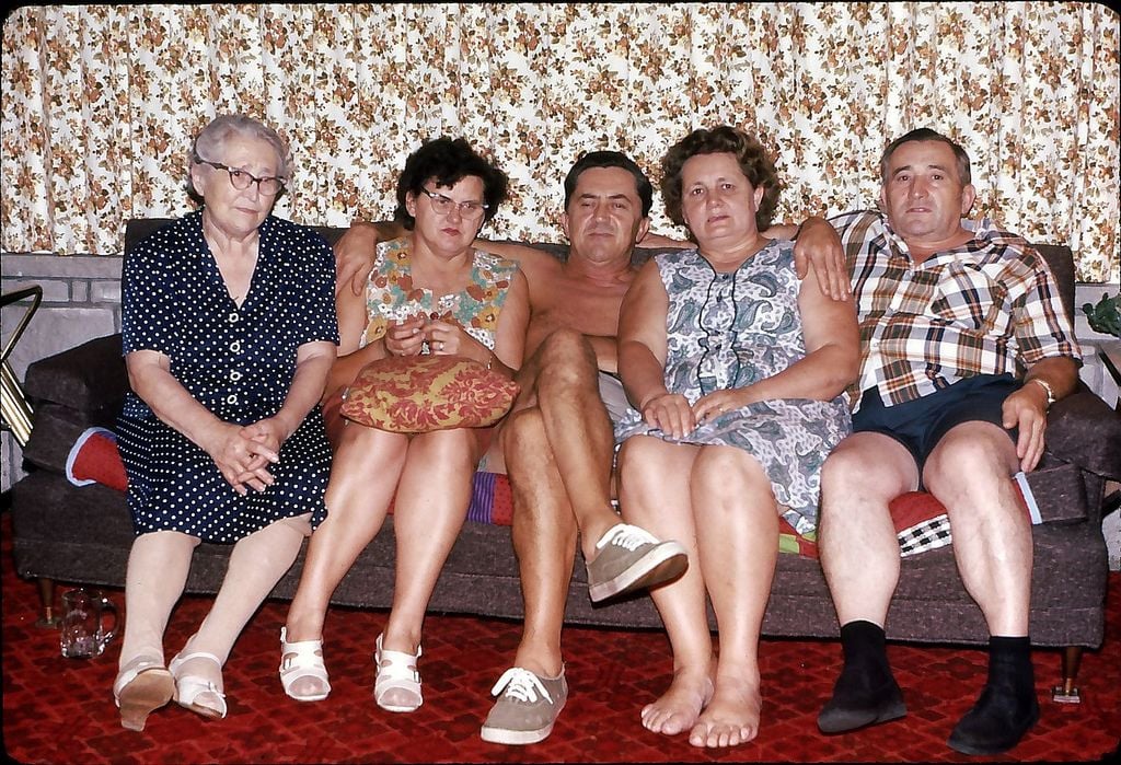 Five adults sit closely together on a couch in a living room with patterned curtains and red carpet, looking at the camera. One person is shirtless, while others wear casual clothing.