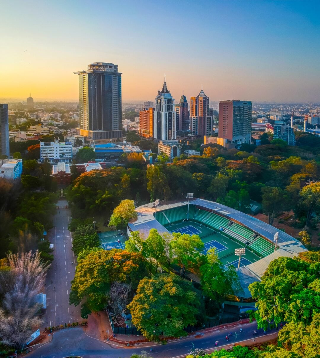 Aerial view of a green stadium surrounded by trees, with tall modern buildings and a cityscape in the background under a clear blue sky at sunset.