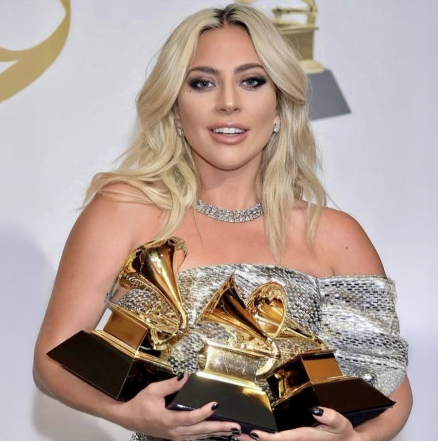 A woman with long blonde hair wearing a silver off-shoulder dress holds three Grammy Awards, smiling at the camera in front of a backdrop with Grammy logos.